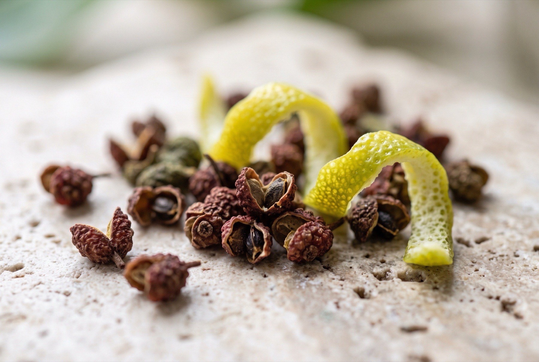 Close-up of cracked Sichuan peppercorns with split reddish husks and a curl of bergamot peel resting on cream travertine stone