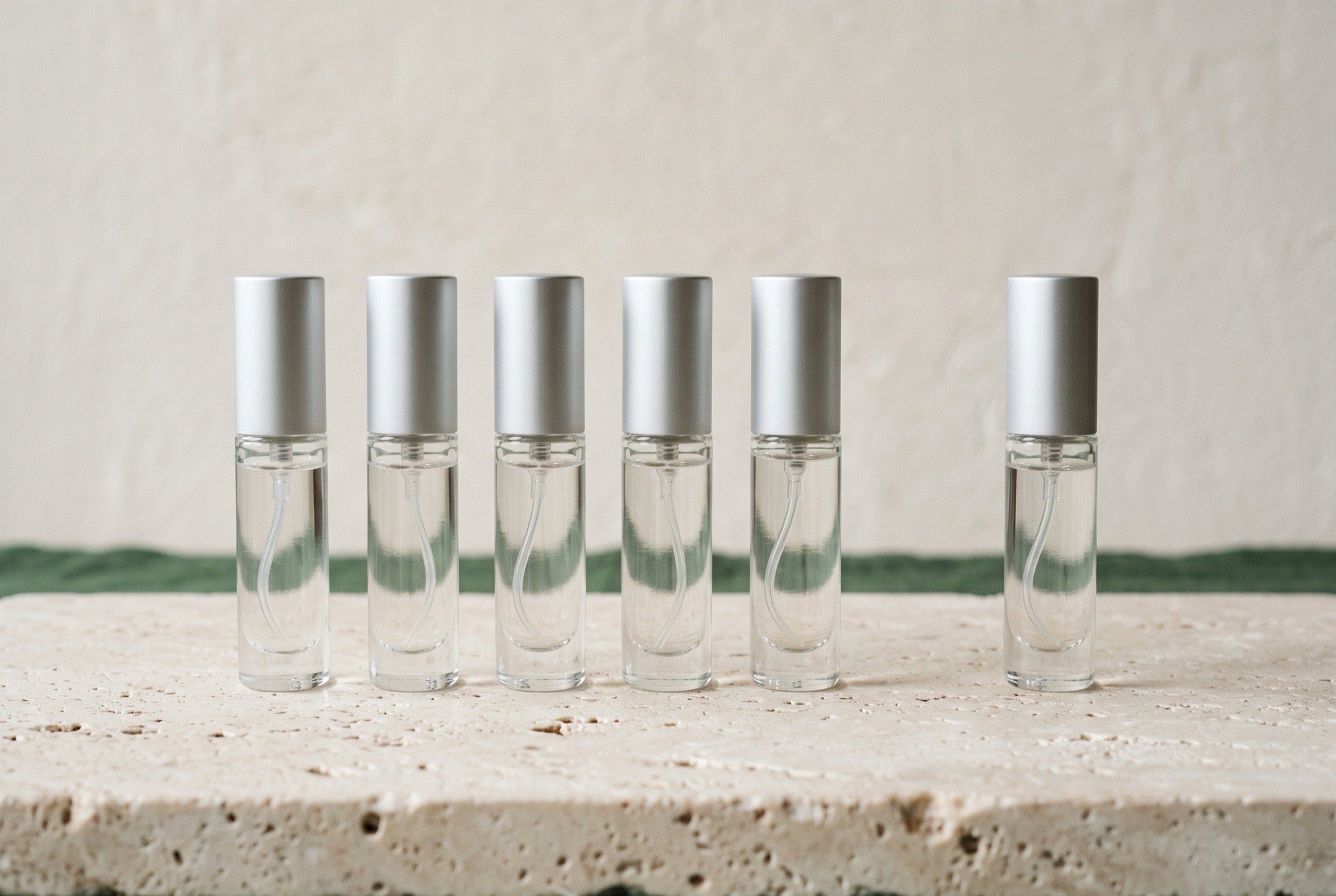 Six generic unbranded clear-glass 5 ml atomizers in a row on cream travertine, one set slightly apart from the other five, with a forest-green linen strip along the lower edge