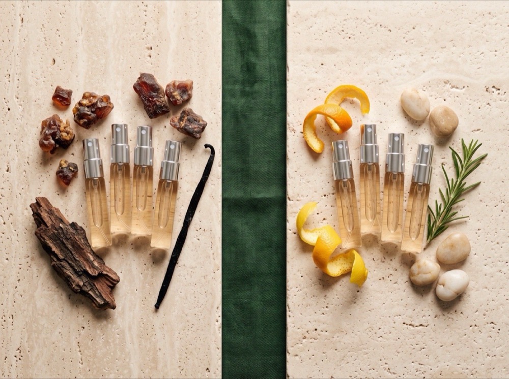 Two groups of identical clear-glass 5 ml atomizers arranged on travertine, one cluster beside dark resinous materials and one beside citrus peel and green herbs