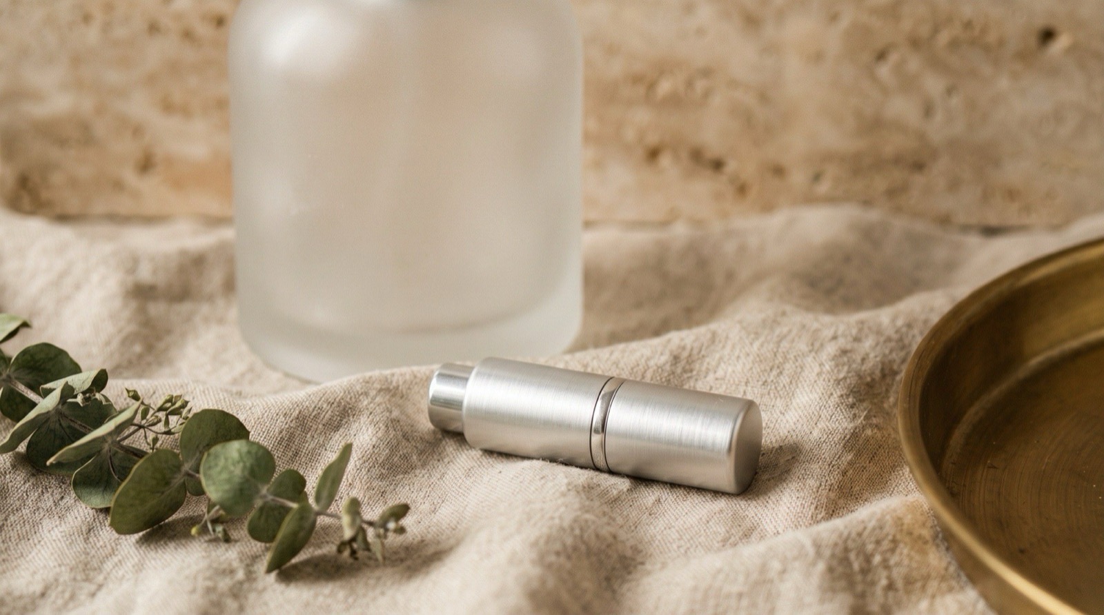 A small opaque aluminium 5 ml travel atomizer lying on linen beside a much larger glass bottle, showing the scale difference